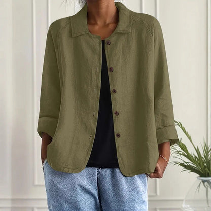 Person wearing an olive green jacket over a black top and blue jeans, standing against a white paneled wall.