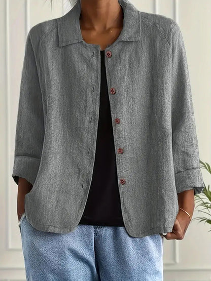 Person wearing a gray button-up shirt over a black top with light blue pants against a white wall.