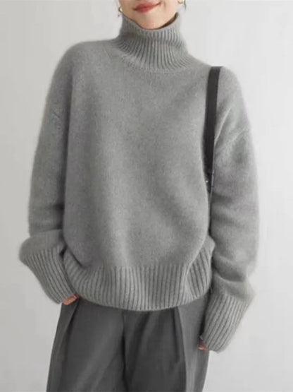 Femme portant un pull gris en laine à col roulé, manches longues, avec un sac noir. Mode automne-hiver, style décontracté et élégant.