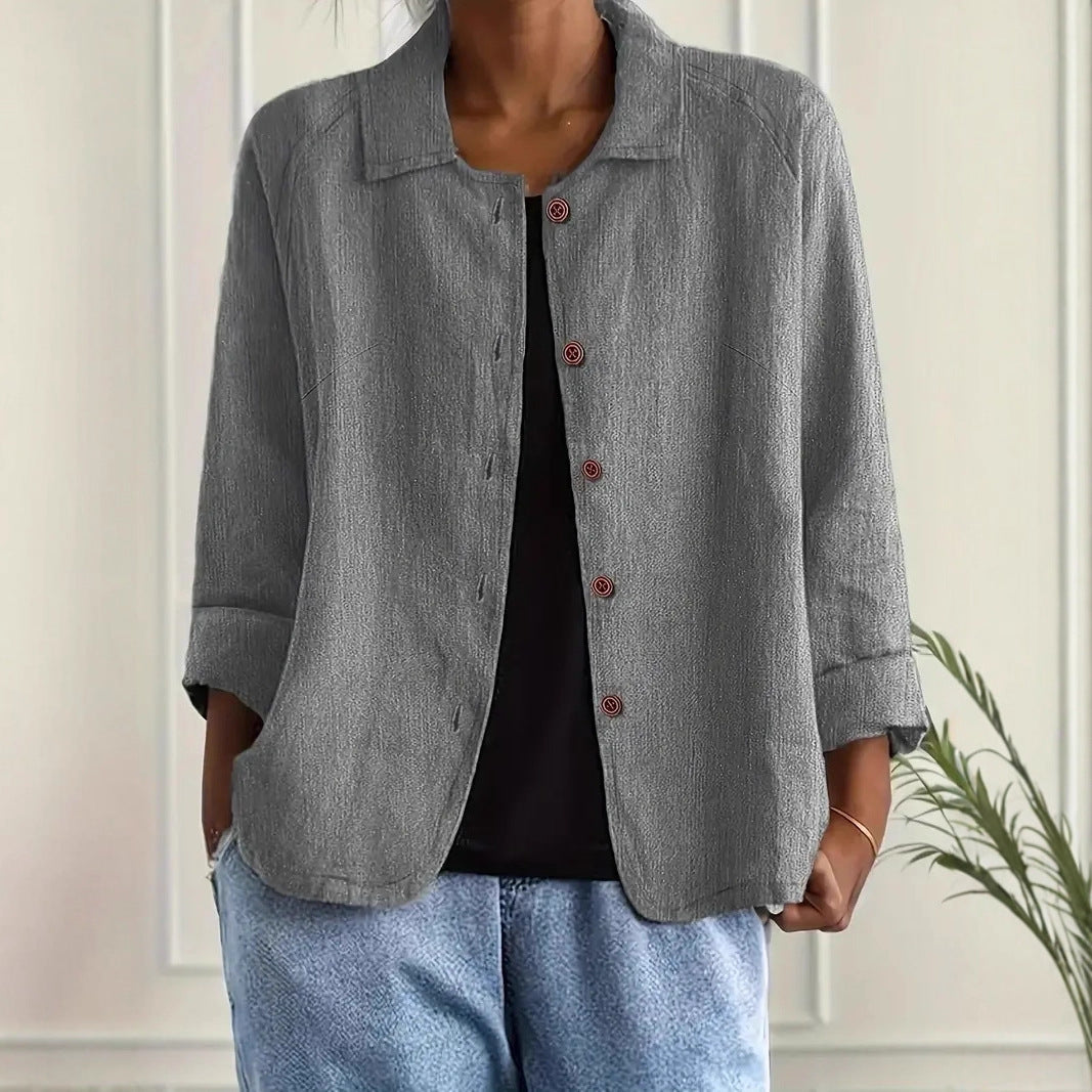 Person wearing a gray jacket over a black top and blue jeans against a white wall.