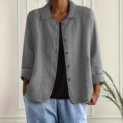 Person wearing a gray jacket over a black top and blue jeans against a white wall.