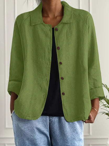 Person wearing a green jacket over a black top and blue jeans against a white wall.