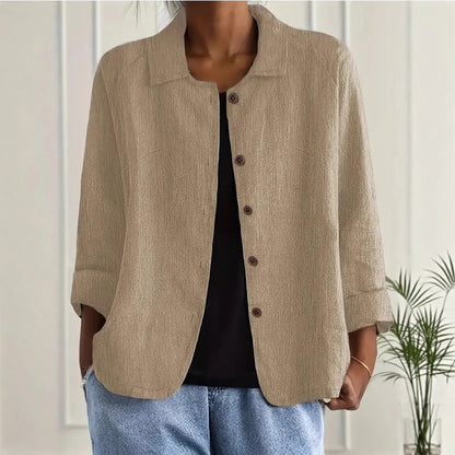 Person wearing a beige jacket over a black top and blue jeans, standing against a white wall with a plant in the background.